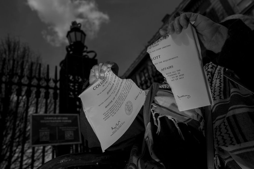 Black and white photo of person wearing a keffiyeh ripping up a Columbia diploma 