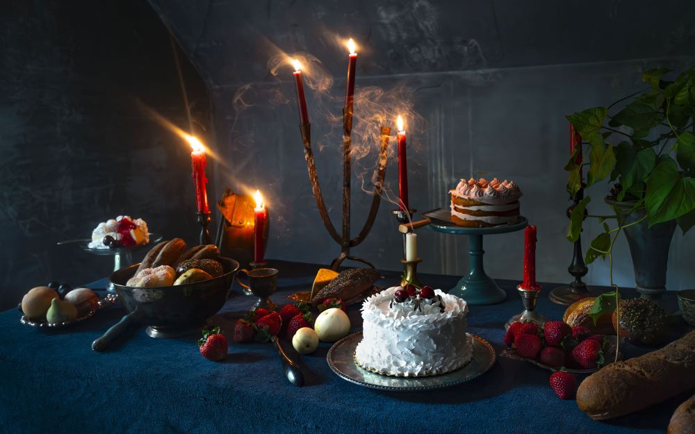 Photo of a cake and fruit spread lit by candles, with deep reds, blues, and cream colors coming through