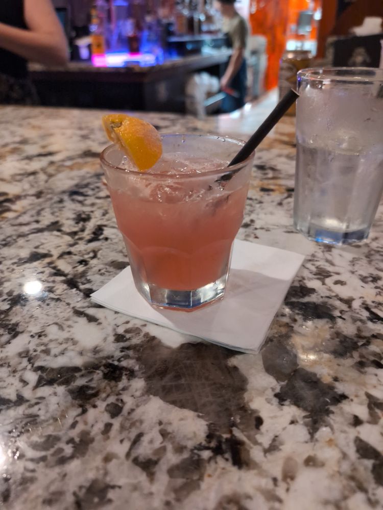 Photo of a drink on a bar. It's called the Midwest Goodbye, it's pink and has an orange slice. It contains tequila, campari, Malört, and sour mix 