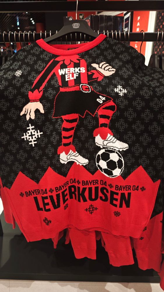 „Schwarzer und roter Weihnachtspullover des Fußballvereins Bayer 04 Leverkusen. Auf der Vorderseite ist eine gezeichnete Fußballfigur mit dem Schriftzug ‚WERKS ELF‘ auf dem Trikot zu sehen, die auf einem Ball steht. Unten steht groß ‚Bayer 04 Leverkusen‘.