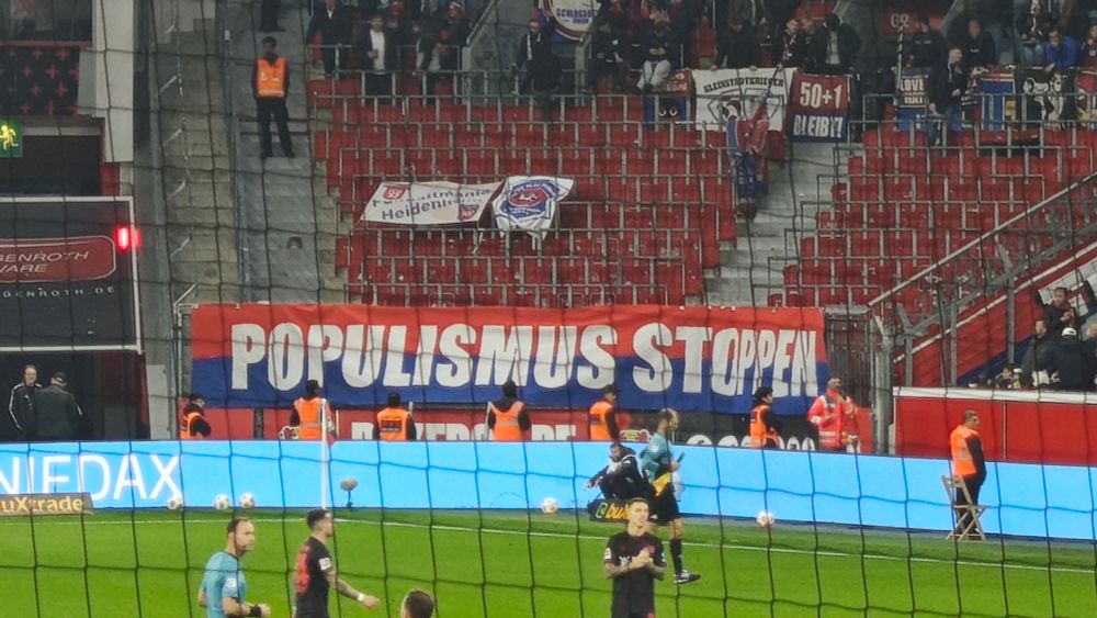 Das Bild zeigt eine Fußballszene in einem Stadion. Im Vordergrund sind Spieler und ein Schiedsrichter auf dem Spielfeld zu sehen, während im Hintergrund Zuschauer auf den Rängen stehen. Auffällig ist ein großes Banner in den Farben Rot, Weiß und Blau mit der Aufschrift „POPULISMUS STOPPEN“, das hinter dem Tor hängt. Mehrere Ordner in orangefarbenen Westen stehen vor dem Banner, und die Tribüne im Hintergrund ist teilweise leer.