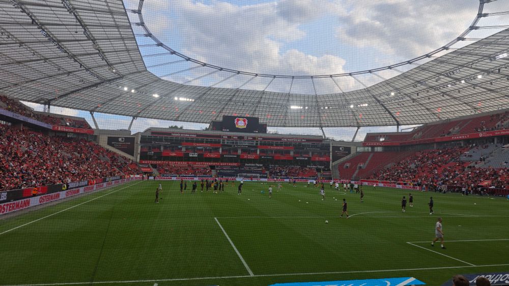 Ein Fußballstadion mit überdachten Tribünen und vielen Zuschauern. In der Mitte des Spielfelds wärmen sich Spieler auf, während das Stadion teilweise gefüllt ist. Auf der großen Anzeigetafel ist das Logo von Bayer 04 Leverkusen zu sehen. Das Stadiondach ist transparent und überspannt die gesamte Arena.

