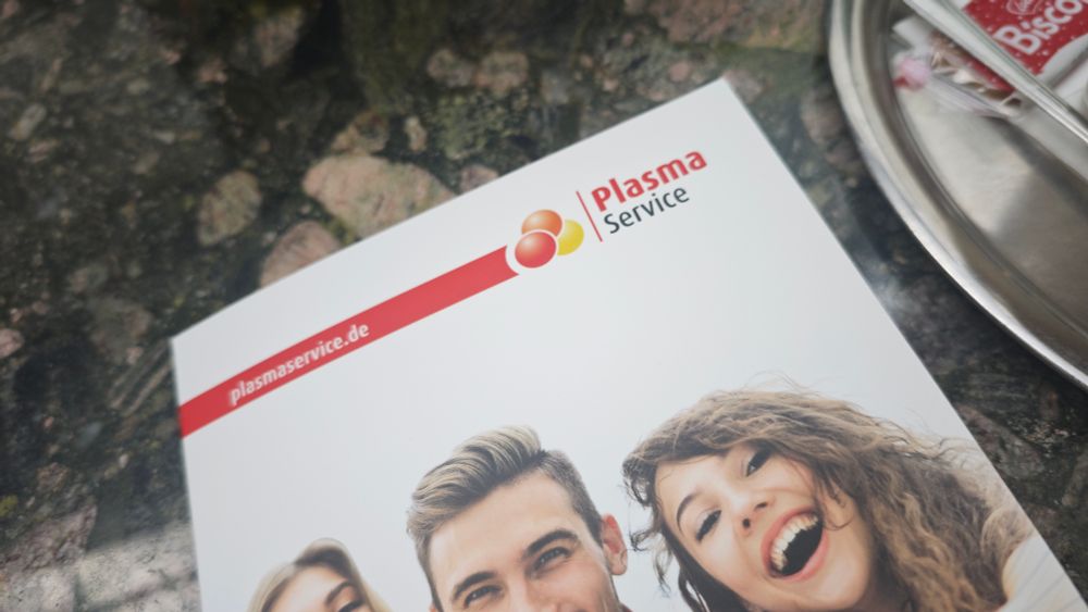 Ein Werbeflyer von "Plasma Service" liegt auf einem Steintisch. Oben auf dem Flyer ist das Logo mit drei farbigen Kreisen (rot, orange, gelb) sowie der Schriftzug "Plasma Service" zu sehen. Darunter lächeln junge Menschen in die Kamera. Auf der rechten Seite des Bildes befindet sich ein silbernes Tablett mit verpackten Biscoff-Keksen.

