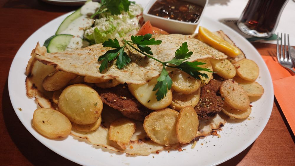 Weißer Teller mit paniertem Schnitzel, gebratenen Kartoffelscheiben, dünnem Pfannkuchen, Petersiliengarnitur, gemischtem Salat mit Gurken und Tomaten, Orangenscheibe sowie einer kleinen Schale dunkler Jägersoße; dahinter ein Getränk auf einem Holztisch.