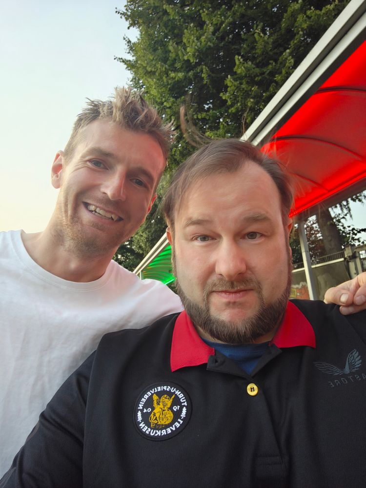 Zwei Männer machen ein Selfie im Freien. Der Mann links trägt ein weißes T-Shirt und lächelt in die Kamera, der Mann rechts trägt ein schwarzes Poloshirt mit rotem Kragen und einem Aufnäher mit der Aufschrift „Turn- und Spielverein Leverkusen von 1904“. Im Hintergrund sind grüne und rote Beleuchtung sowie Bäume zu sehen.