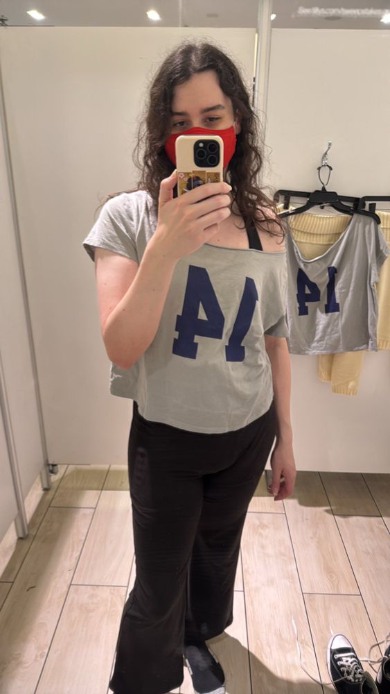 A photo of Eve (TheMoxyFoxy) wearing a light grey off-the-shoulder shirt with the number “14” on it in blue (reversed due to camera) and black leggings in a dressing room