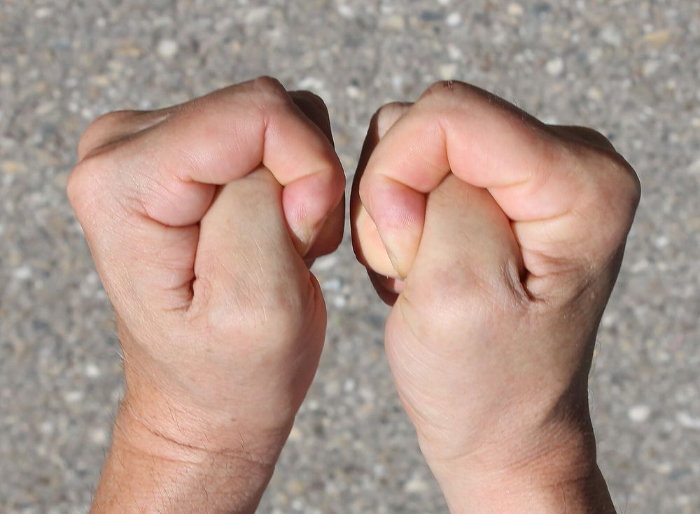 Gedrückte Daumen an zwei mittelalten, von der Behaarung her wohl männlich gelesenen Händen vor einem Steinbodenhintergrund
