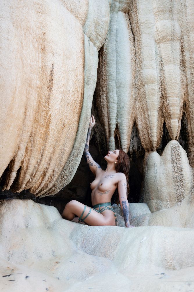 A woman poses in a mineral cave surrounded by dramatic beige, cream, and pale teal formations that echo the colours of her teal and gold harness. The textures of the cave walls are soft and rippled, shaped by time and water, creating a surreal, organic backdrop. Kneeling gracefully on a smooth mineral surface, she reaches upward with one arm, her gaze following in a gesture of quiet awe. Natural light filters through the scene, casting gentle highlights that enhance the harmony between her pose, the harness, and the mineral tones, evoking a sense of and otherworldly reverence.

