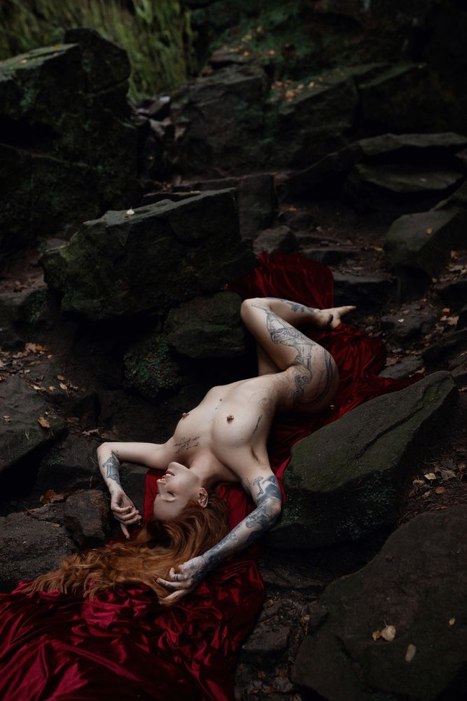An artistic photograph depicting a nude woman reclining on a sloped pathway of jagged, sharp stones. Her body arches over the path, with her head tilted downward, allowing her long, cascading red hair to flow dramatically like liquid fire. Beneath her, a rich velvet fabric, deep crimson in colour, unfurls and spreads across the stones, resembling a river of blood. The composition evokes strong biblical and mythological themes, suggesting imagery of the underworld and Dante Alighieri’s Inferno. The stark contrast between her pale skin, fiery hair, and the dark, textured background enhances the scene’s haunting and otherworldly atmosphere