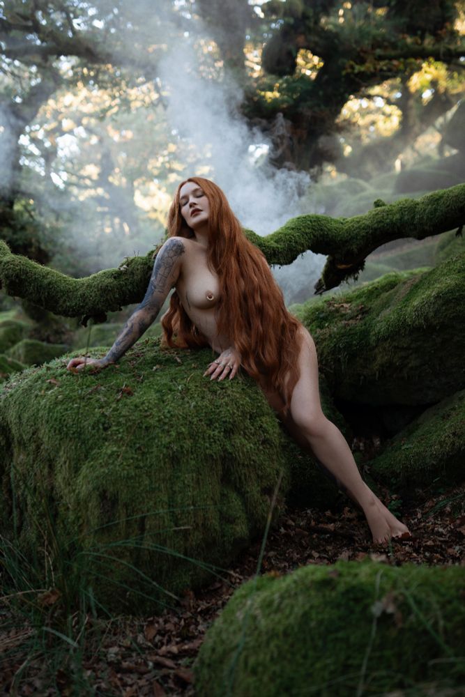 A nude woman with long flowing red hair reclines sensually on a moss-covered boulder in an enchanted forest. Her arm rests against the rock, her head tilted back with a dreamy, almost spellbound expression. Mist rises in the background, curling through the twisted tree branches and creating an ethereal, otherworldly atmosphere. The scene feels mythological, evoking imagery of forest nymphs, Pre-Raphaelite muses, and ancient enchantresses.