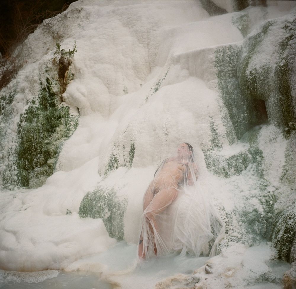 This is a medium-format colour photograph depicting a figure reclining against a natural white and green mineral backdrop, evoking a dreamlike and surreal quality. The composition draws strong references to 18th-century veiled sculptures, such as Giuseppe Sansevero's Cristo Velato and Antonio Corradini's Vestal Virgin. The bare skin of the figure is faintly visible beneath a delicate, semi-transparent fabric, mirroring the masterful marble sculpting techniques that created the illusion of flesh beneath veils. 

The interplay between the sheer fabric and the natural textures creates a hauntingly ethereal atmosphere. The muted colours of the scene - soft whites with pale green moss-like formations - further emphasize the otherworldly, serene, and spectral aesthetic of the image. The photograph captures an evocative blend of fragility, mystery, and timelessness.