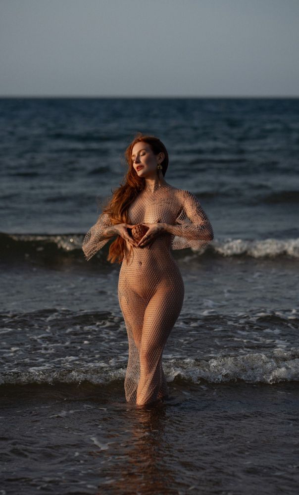 This photograph draws inspiration from the myth of Venus's birth, the Roman goddess of love and beauty, who is said to have risen from the seafoam. The model stands poised in the ocean, embodying the mythical essence of emergence and rebirth. She wears a see-through net dress, a deliberate nod to the sea as it resembles fishing nets, connecting her to maritime symbolism. A seashell, shaped like an anatomical heart, is held gently over her chest, representing life, love, and emotion. The interplay of warm, golden tones from her hair and the shell against the deep blues of the ocean creates a striking contrast, emphasizing the serene yet powerful energy of the scene.