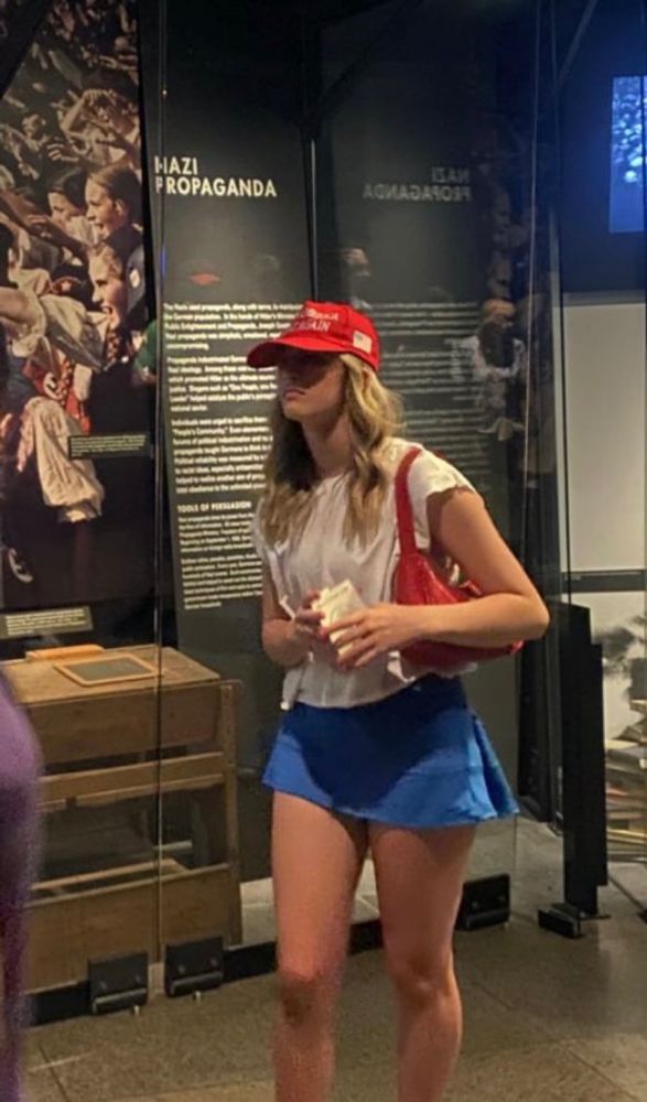 Girl with a MAGA hat standing in front of a museum exhibit on Nazi propaganda.
