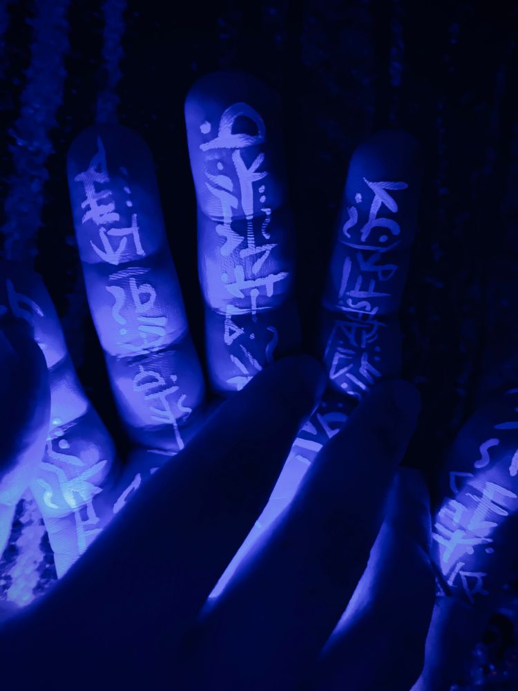 White glyphs painted on inner palm of hand under blacklight. Second hand lays over it partially blocking the light.