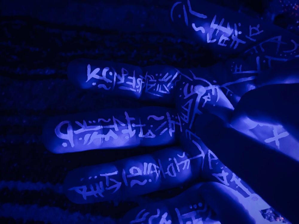 White glyphs painted on inner palm of hand under blacklight. Second hand lays over it partially blocking the light.