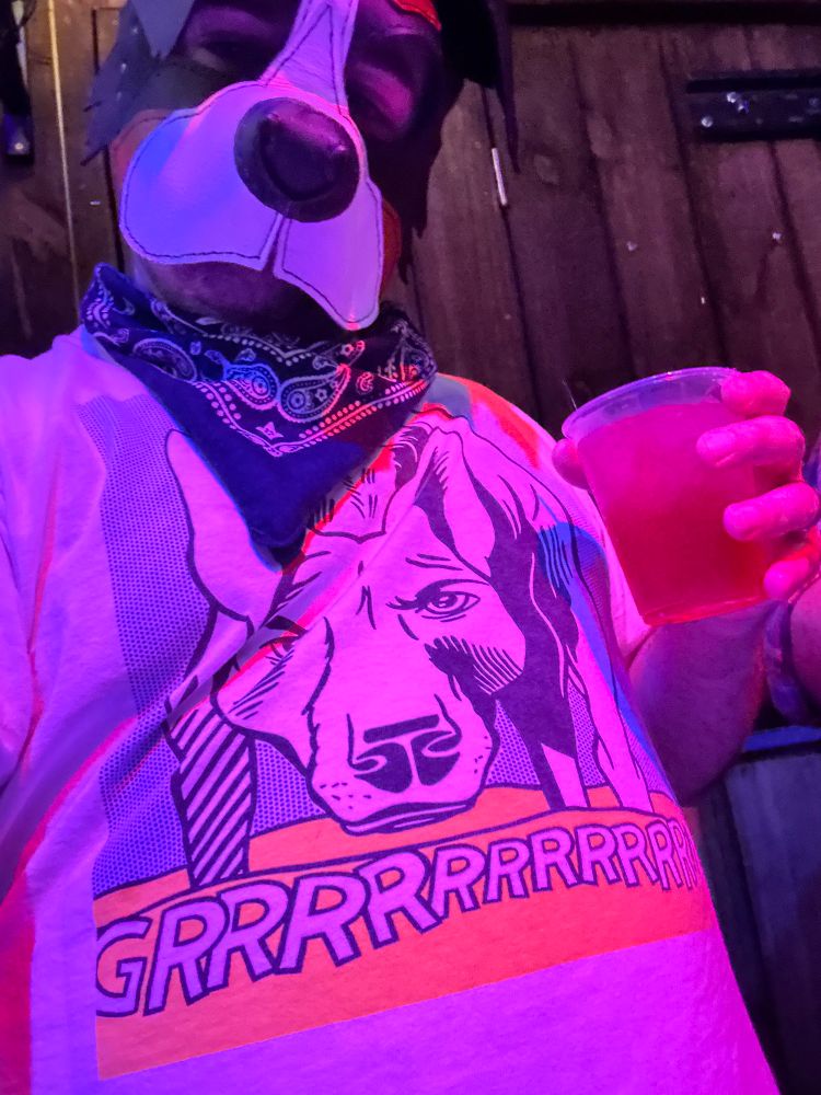 Selfie of me in pup hood, cast in pink light wearing a pop art tee of a dog saying GRRRRR! and navy paisley bandana holding a drink.