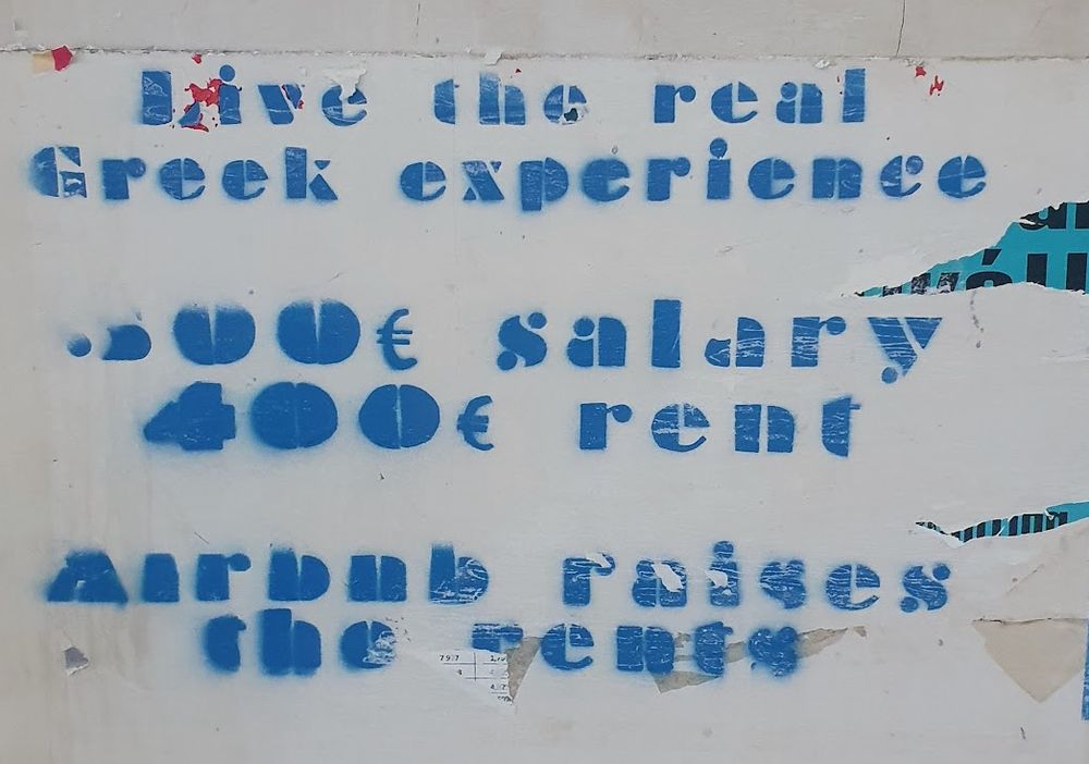Grafitti frå Athen. Blå skrift på kvit vegg: 
Live the real Greek experience. 500€ salary, 400€ rent. AirBnB raises the rents.