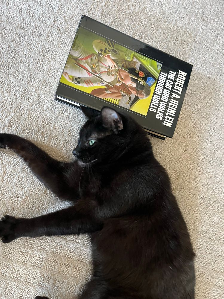 My black cat hanging out next to a copy of Heinlein’s “The Cat Who Walks Through Walls”