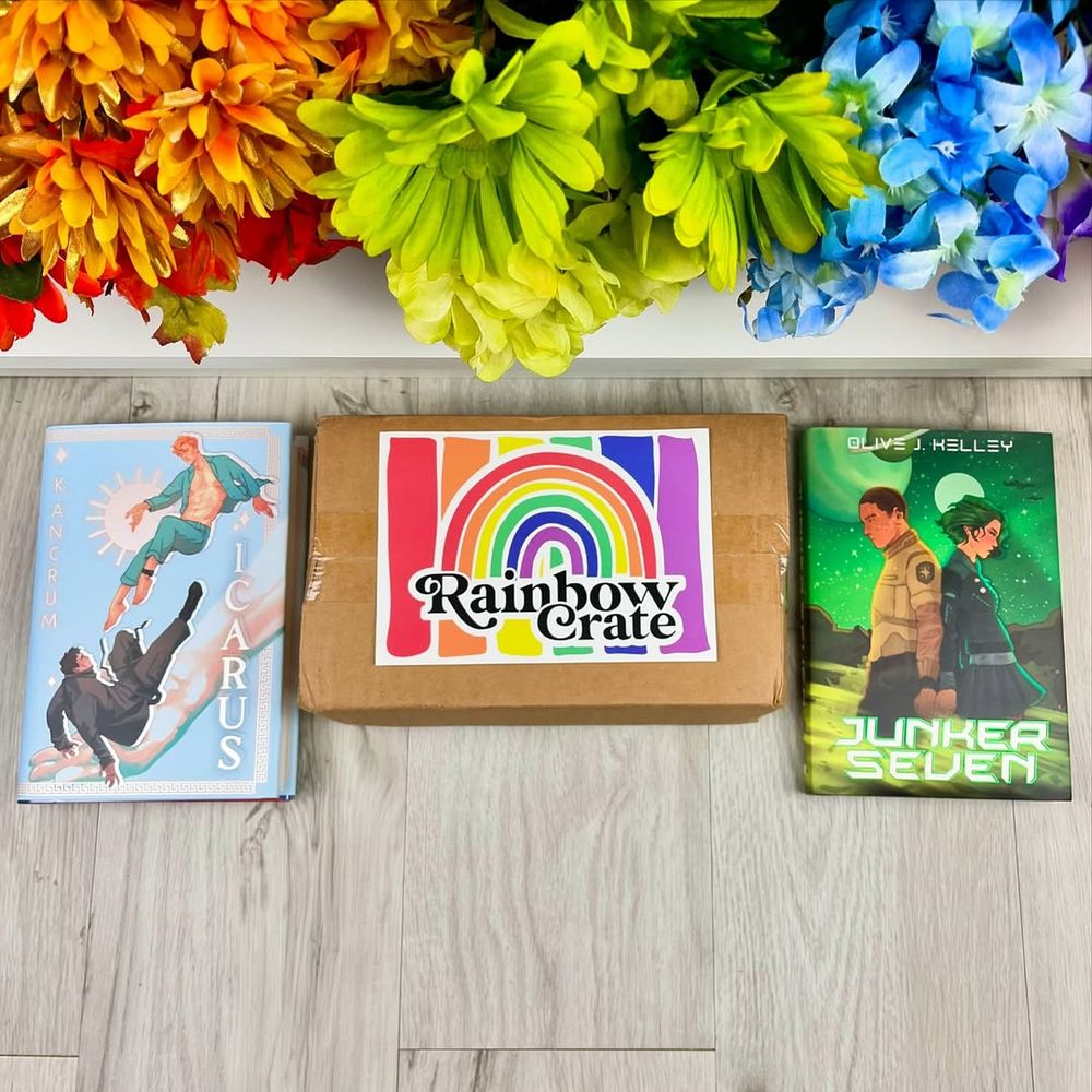 A top down photo of Icarus, a brown Rainbow Crate box and Junker Seven. The books sit on either side of the box. Above are a rainbow of flowers. Everything sits on a gray wooden floor. 