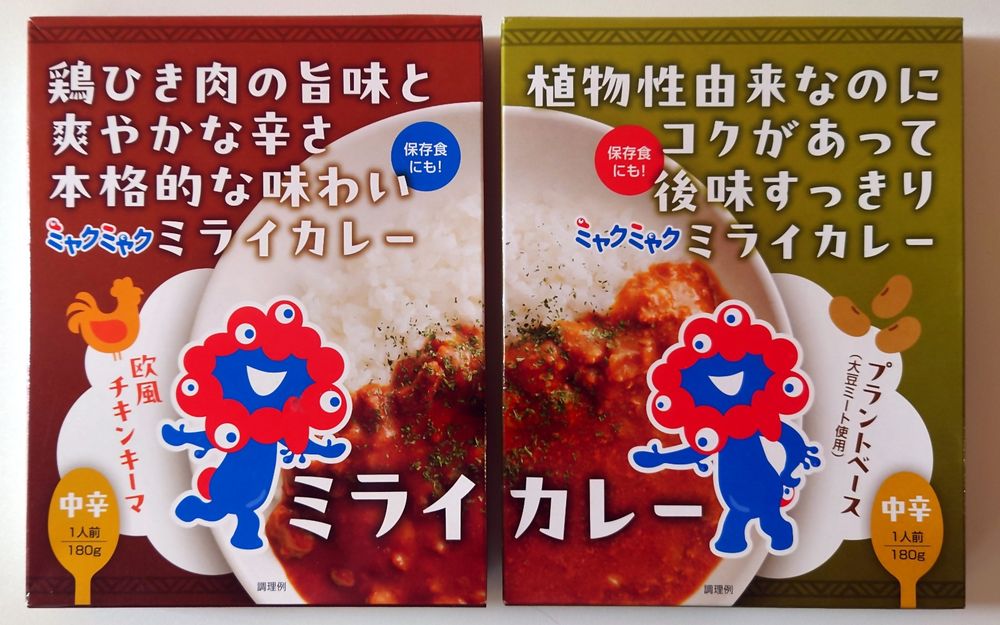 Two packs of curry of the future. Brown chicken pack on the left, green vegan pack on the right. The blue and red character with many eyes is the official mascot of the 2025 World Expo.