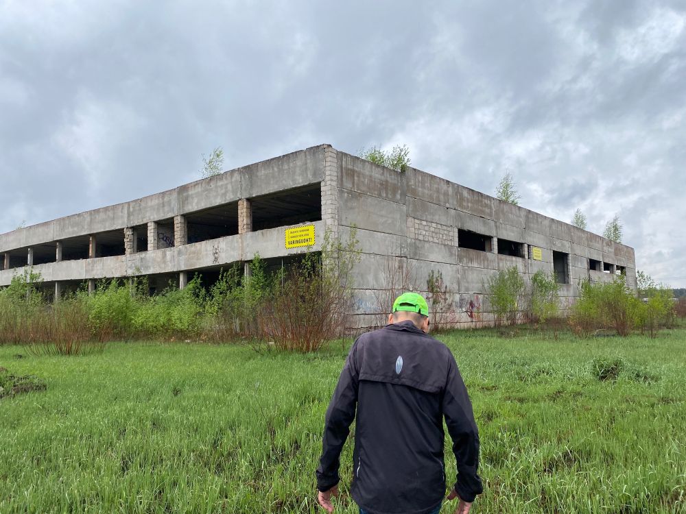 A man in a green baseball cap and a black coat walks across grass towards the shell of an abandoned two-storey building. There’s a yellow warning sign on the building stating that the place is unsafe