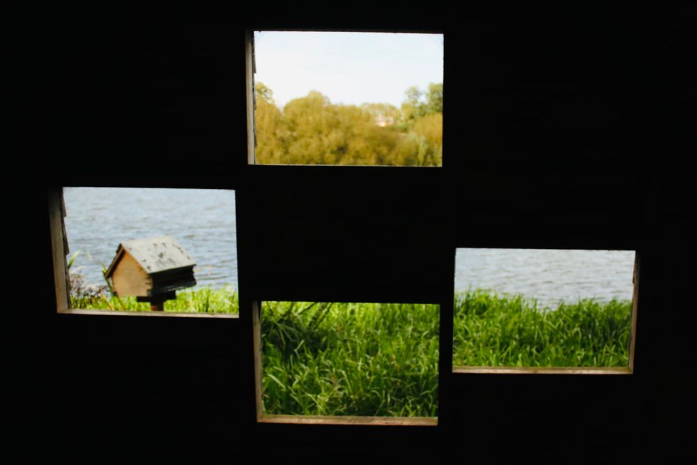 Four openings set in a diagonal pattern showing the scene beyond a bird hide. One shows sky and treetops, another shows water and grass, the third just grass, and the fourth frames a bird feeder shaped like a miniature house