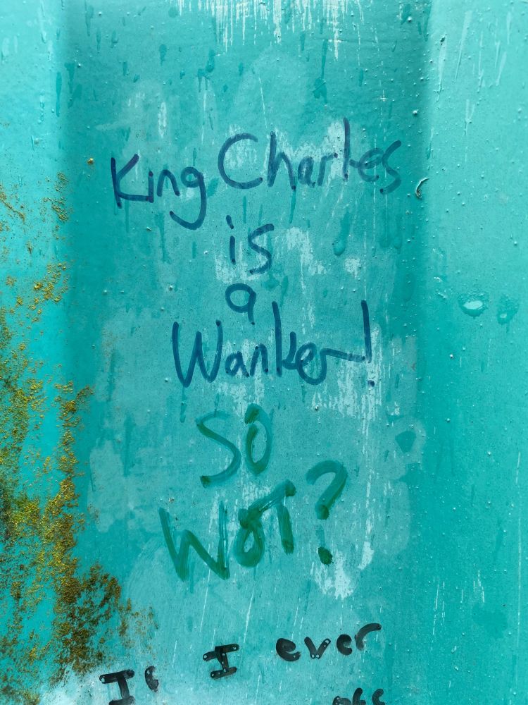 Close up of graffiti on the blue corrugated iron of the bridge. It reads ‘King Charles is a Wanker!’ And beneath that someone else has written ‘So Wot?’