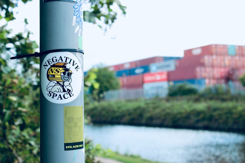 A sticker on a lamppost that reads ‘Negative Space’. Another beneath it shows a skull with the words Evil Acid Cult. In the soft focus background there’s a canal and a stack of shipping containers