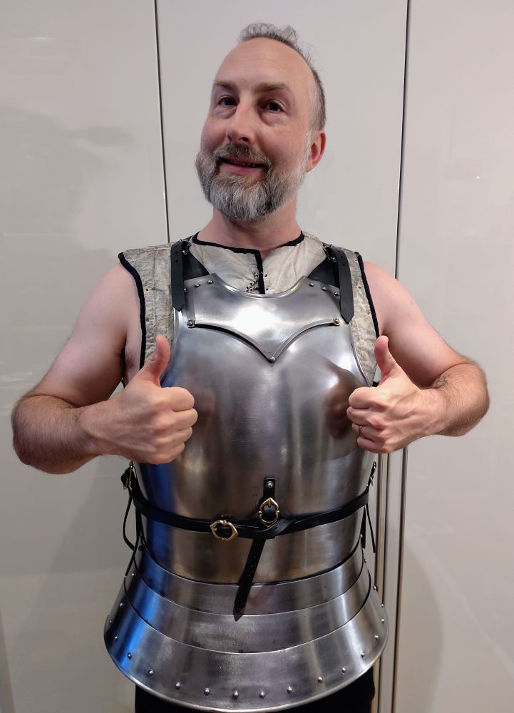 Ein nicht mehr ganz junger Mann steht vor einem weißen Hintergrund. Er trägt eine glänzende Brustplatte in historischem Stil. Darunter trägt er eine gepolsterte Weste. Der Mann lächelt und zeigt mit zwei Daumen nach oben.