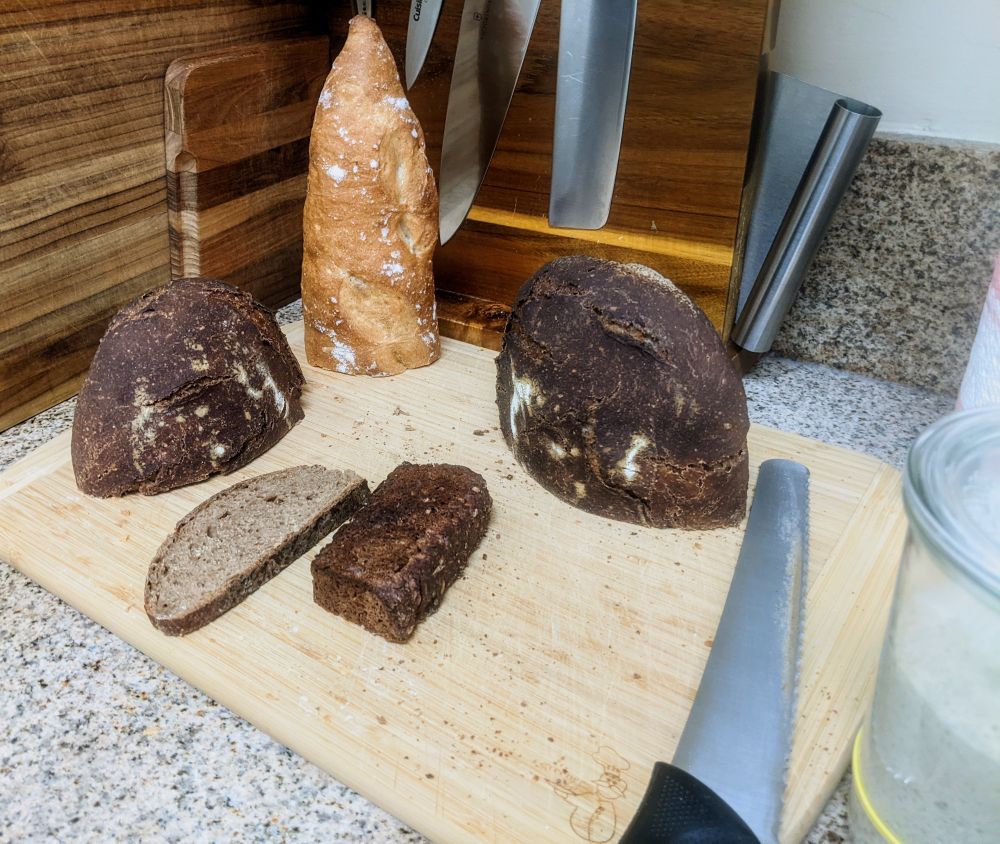 Breads on a cutting board, a bread knife to the side and a jar with a sourdough starter at the edge of the frame.