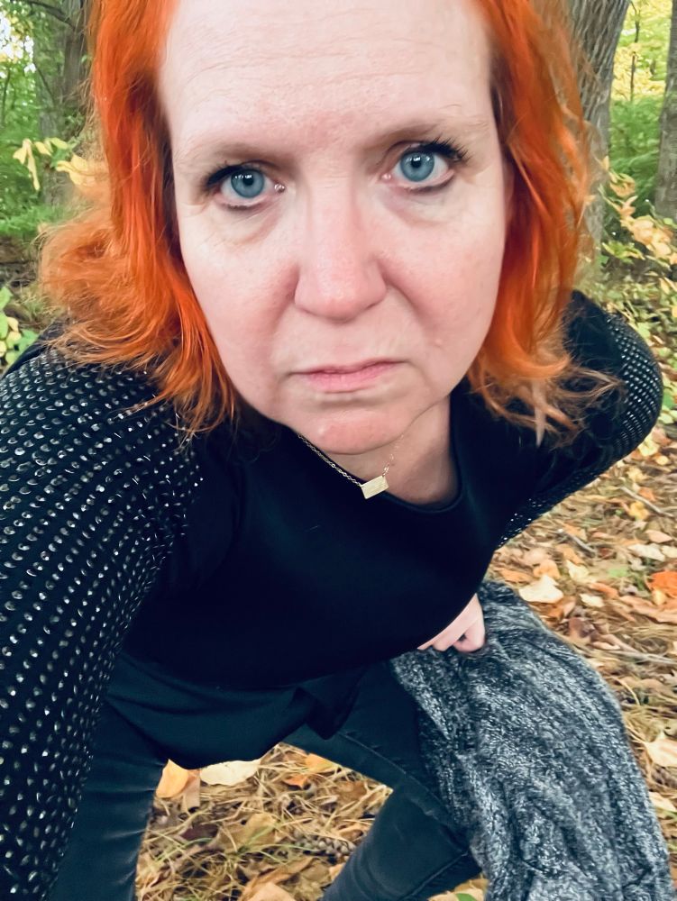 Anika poses close to the camera to show off her sleeves set to look like chain mail, black peplum top and silver cardigan bunched in her hands. She has bright copper hair and is wearing a small gold necklace with a rectangle pendant.