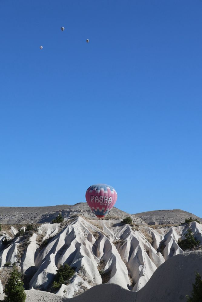 A red white and blue hot air balloon rising from an eroded white tufa rock valley  into a blue sky with three other distant balloons in the Kapodokya region of Turkey. 