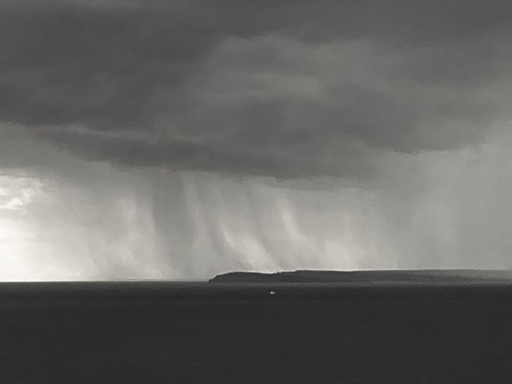 A black and white photograph of a small white boat on the sea in a passing shower between Orkney and the Scottish mainland’s most northerly point at Dunnet Head. 