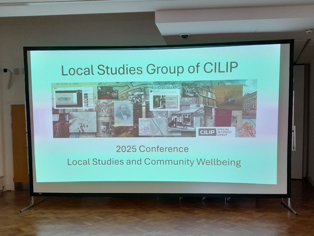Large projector screen showing home slide from PowerPoint presentation for the Local Studies Group of CILIP 2025 conference. The conference theme is Local Studies and Community Wellbeing.