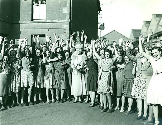 A crowd of cheering women workers at Hadens factory in Trowbridge stand either side of Queen Mary who visited the factory during the Second World War. In the background three women look out from a first floor window. Queen Mary is wearing a long coat with a fir lined collar and is wearing a hat. 