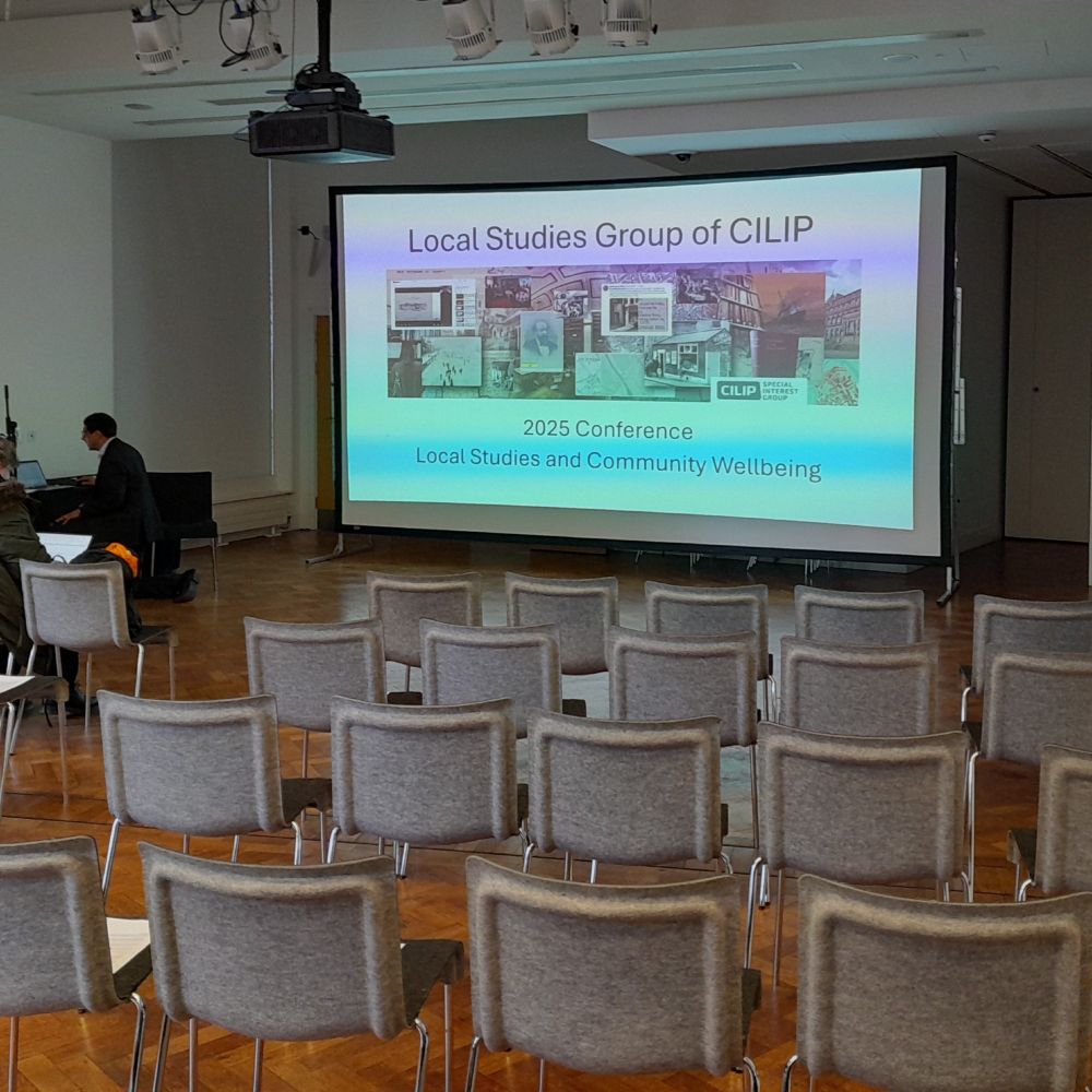 View from the back of a hall showing chairs, digital projector and screen plus one person to the left of the image setting up the PowerPoint presentation on a laptop. The screen shows the home slide for the Local Studies Group of CILIP conference on Community & Wellbeing.