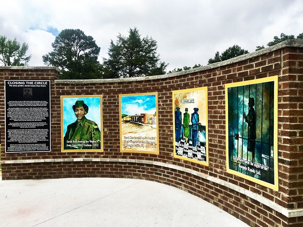 Monument with curved brick wall, painted pictures, and text engraved on a plaque. Trees and cloudy sky in the background.