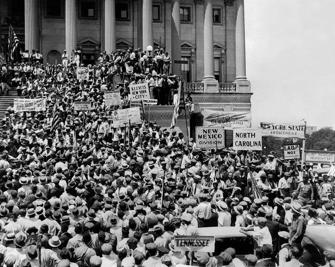 Black and white photo of crowd (most are wearing hats) facing building with columns, carrying signs naming states. 