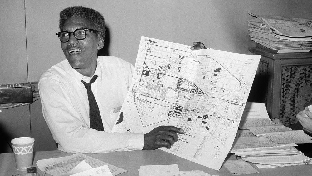 Bayard Rustin with plans for March on Washington. Black and white photo -- man wearing tie, with glasses, pointing to a map, sitting at a table with papers and a paper cup. 