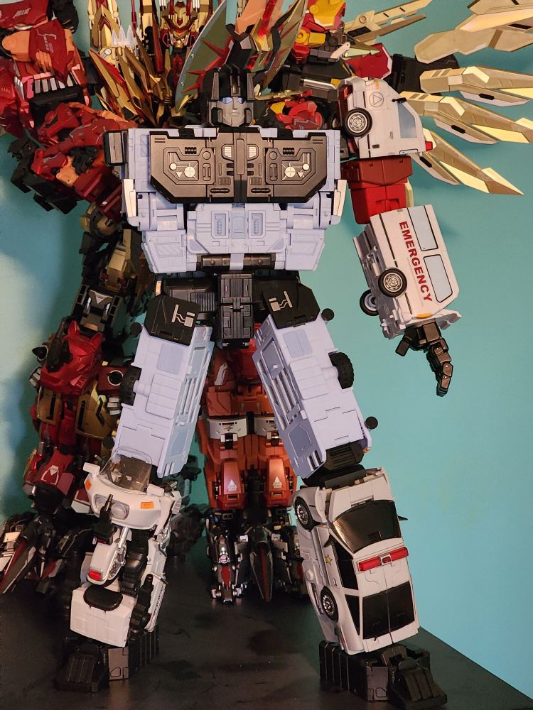 Blue and grey robot made from four emergency vehicles missing it's right arm. Left arm is a white and red ambulance. Left leg is a grey police car. Right left is a white police motorcycle.