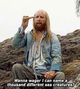 A balding pirate with long blond hair and a beard in a blue coat and baggy pants whittling. The text at the bottom says "Wanna wager I can name a thousand different sea creatures?"