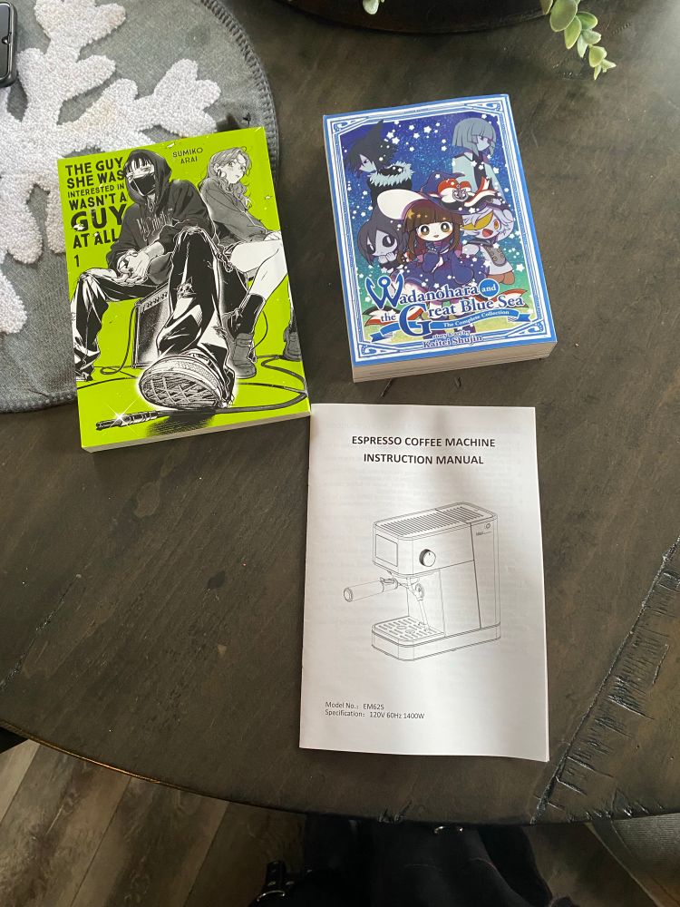 Three books lay on a table: The Guy She Was Interested in Wasn’t a Guy at All (Volume 1), Wadanohara and the Great Blue Sea (Complete Collection), and a Espresso Coffee Machine manual.
