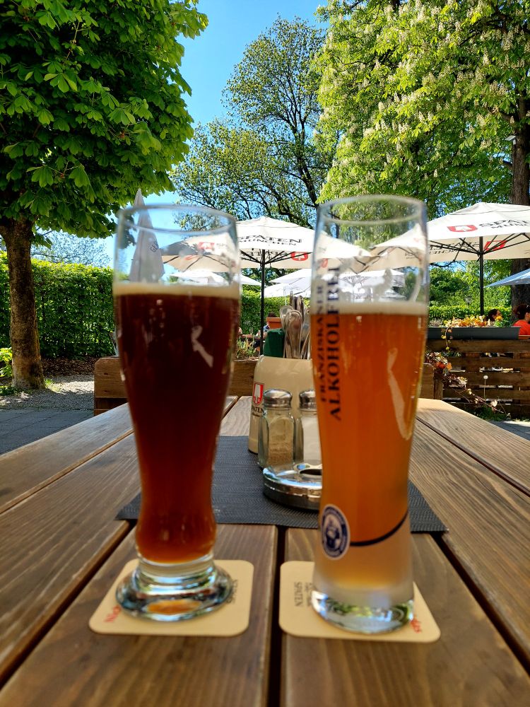 Zwei volle Biergläser im Hintergrund blauer Himmel und grüne Laubbäume im Biergarten.  