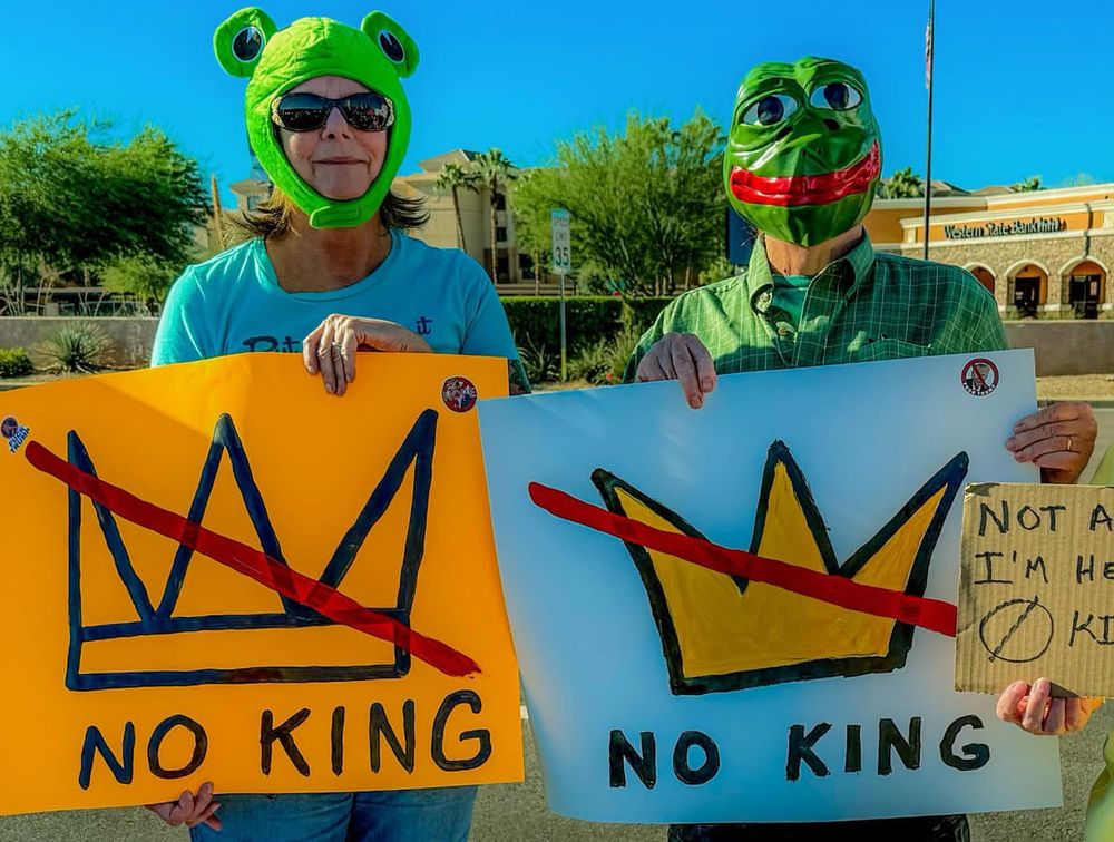 A woman with frog headgear holding a No King sign with a crossed out crown. She’s standing next to another person who has a frog head on  and also holding a No King sign