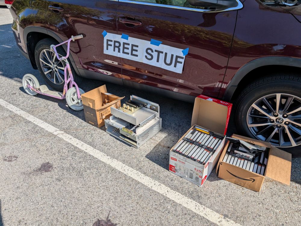 A pile of things including an electronics case, a small child's scooter, and two boxes of VHS tapes. Taped to the car door is a sign that reads "FREE STUF"