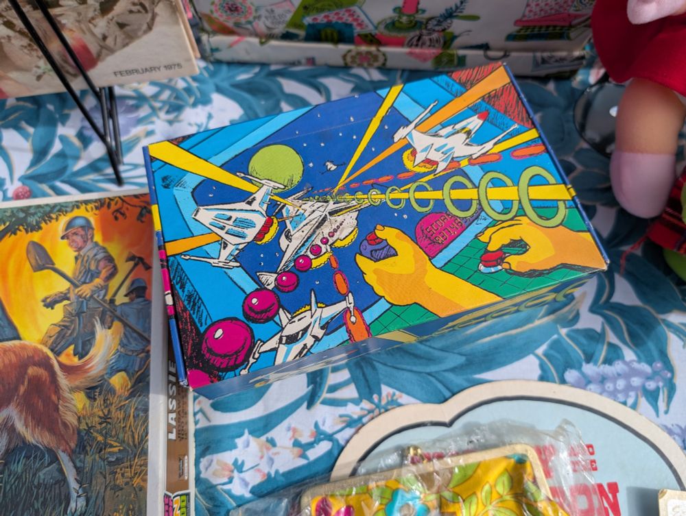 A child's cardboard pencil case that depicts a space arcade game with ships and projectiles flying out of the screen. In the foreground you can see two hands, one on a joystick and the other on a fire button. There's a score counter in the lower right of the screen.
