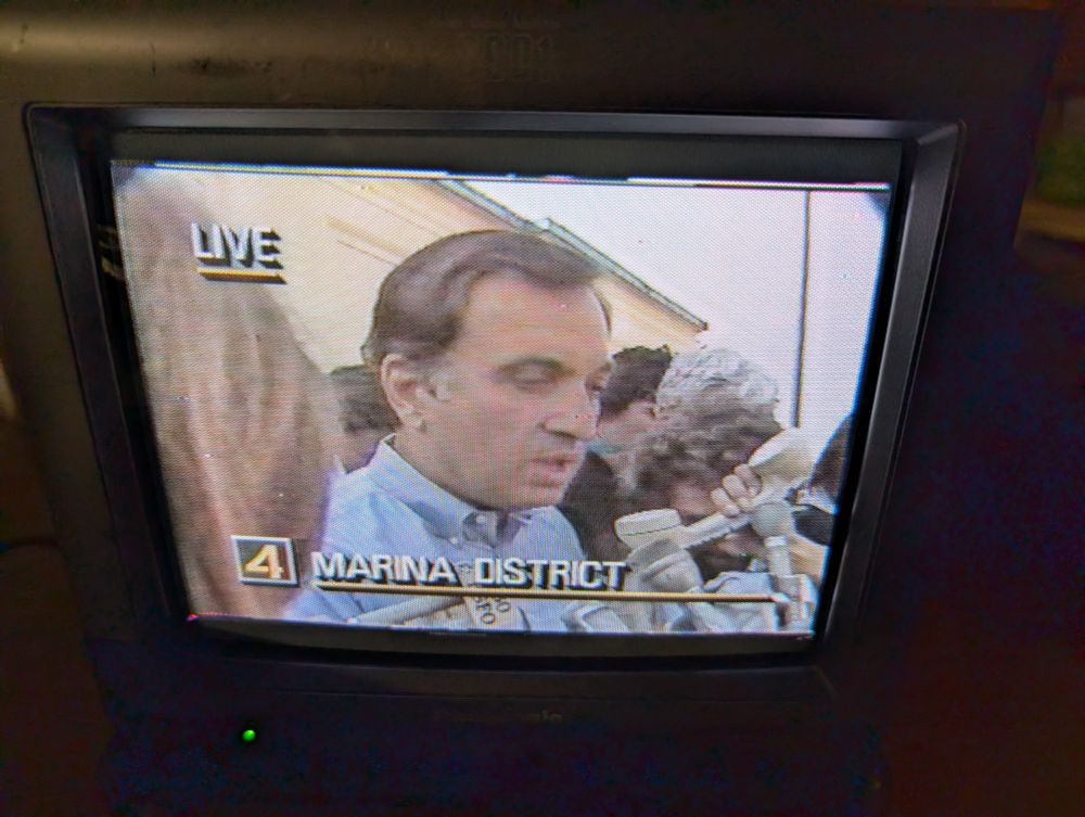 Another screenshot of a news broadcast. A person is giving a press briefing. Bottom graphic says 4 Marina District. One of the many mics in front of him is a beige handset from an analog phone