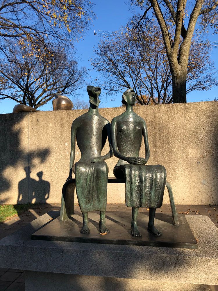 Bronze sculpture of stylized man and woman sitting next to each other on a bench. It is outside with bare trees visible in the background.