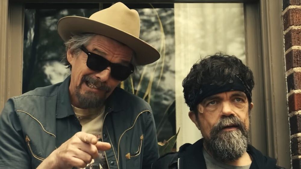 Actors Ethan Hawke and Peter Dinklage from the TV show The Lowdown. Ethan is wearing a cowboy hat, sunglasses and a blue denim western shirt. He's pointing and talking out of the side of his face. Dinklage has a black headband and a scraggly beard.