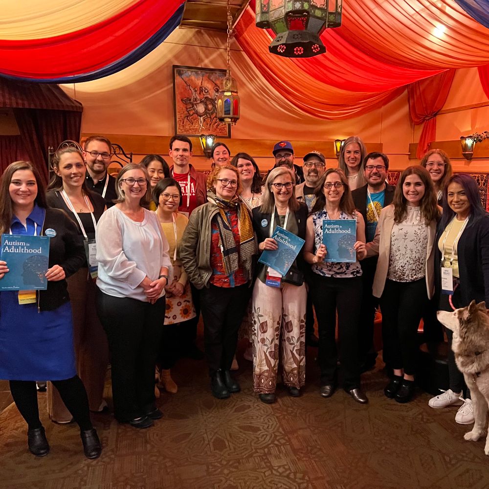 Group photo of 19 people and a service dog posing in a Moroccan restaurant. Most of the people are Autism in Adulthood Editorial Board Members. 